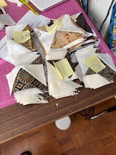 Wanderlust-Sharon Several stacks of small fabric triangles, each pile labeled with a yellow sticky note and number, are arranged on a wooden table next to a pink cutting mat and some sewing supplies.