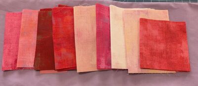 Wanderlust-SueG-FQ Rectangular fabric pieces in shades of red, pink, and peach are arranged in a row on a lavender background. The fabrics have a textured, slightly mottled appearance.