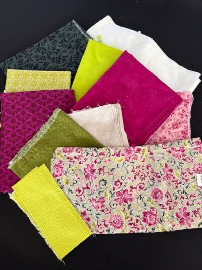 Wanderlust-SueI An assortment of colorful fabric pieces, including floral prints and solid colors such as pink, yellow, green, white, and black, laid out on a dark surface.