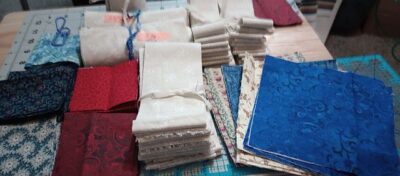 Wanderlust-WendyZ Stacks of folded fabric in various colors and patterns, including blue, red, cream, and floral designs, are arranged on a cutting mat on a wooden table, ready for sewing or quilting projects.