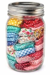 A mason jar filled with neatly folded, colorful patterned fabric squares and a pair of small gold scissors. The fabrics feature polka dots, florals, and checks in various bright colors.