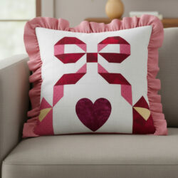 Square throw pillow with pink ruffled edges on a beige couch. The pillow features a patchwork design of a red and pink bow and a heart, with yellow accents, creating a whimsical, cozy look.