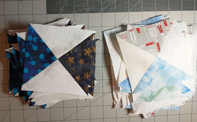 wanderlust-Carole-QST Two stacks of quilt blocks on a cutting mat: one stack features blue and white triangle patterns, some with stars or polka dots; the other stack shows blocks with pale blue, grey, and red triangle designs.