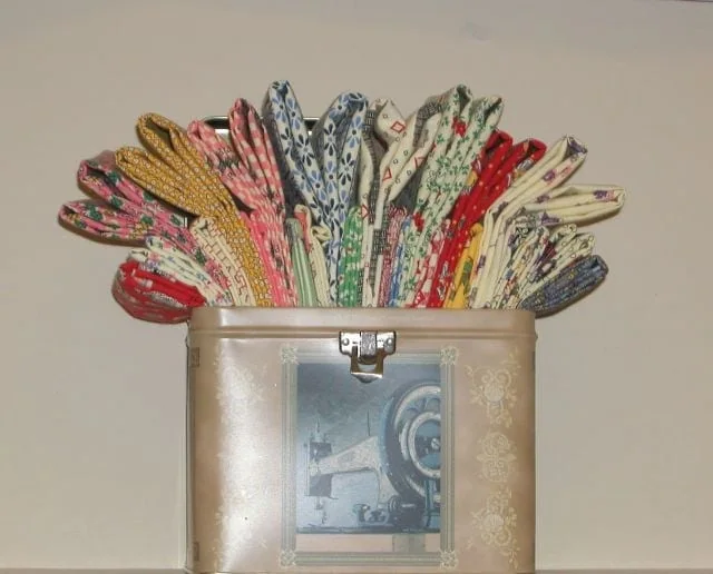 Box-of-FQ A beige tin box decorated with a vintage sewing machine image holds neatly folded, colorful patterned fabrics standing upright and fanned out at the top.