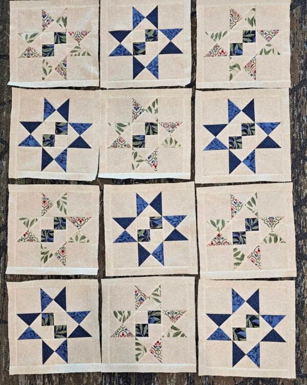 Twelve quilt blocks arranged in rows, each featuring a central pattern of small squares and star shapes made with blue, green, and floral fabrics on a beige background. The blocks rest on a textured dark surface.