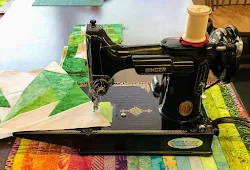 A vintage black Singer sewing machine sits on a table with a colorful patchwork quilt under its needle and a spool of white thread on top.