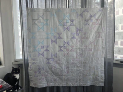 Khai-Baby-Quilt A black and white cat sits on a windowsill next to a hanging quilt with pastel star patterns, displayed in front of sheer curtains with city buildings visible through the window.