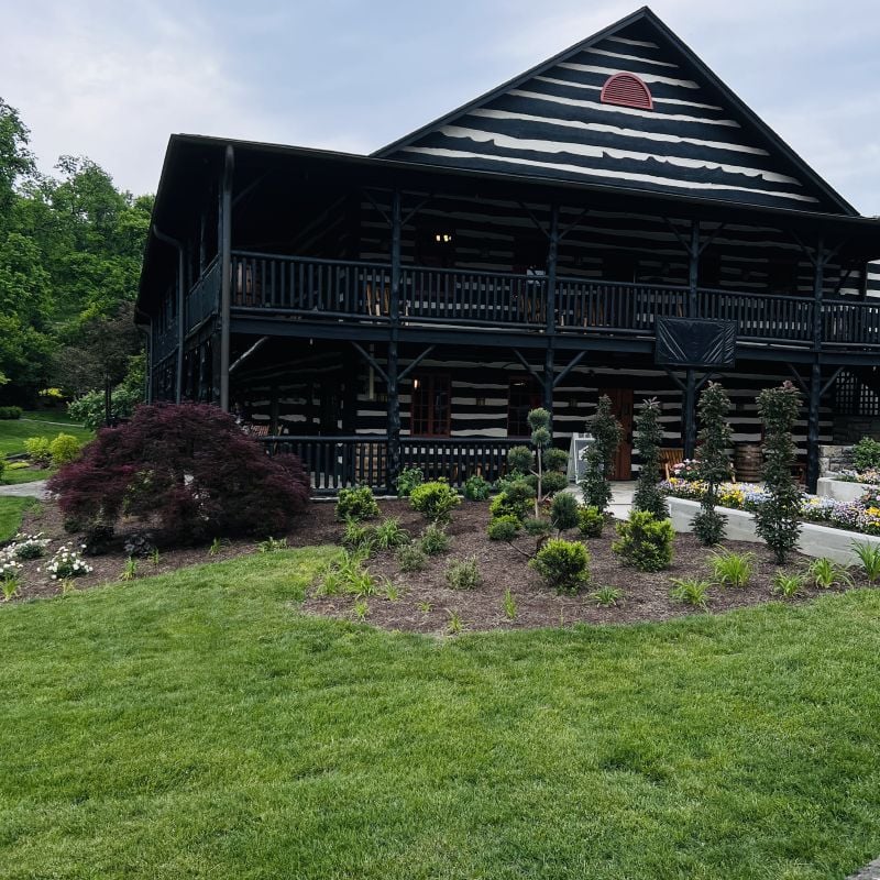 A large black-and-white log cabin with a wraparound porch is surrounded by manicured lawns, shrubs, and landscaped garden beds, set against a backdrop of green trees under a cloudy sky.