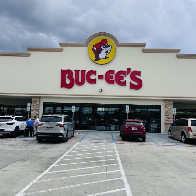 The entrance of a Buc-ee’s convenience store with its large red logo and beaver mascot above the doors. Several cars are parked in front, and a few people are near the entrance under a cloudy sky.