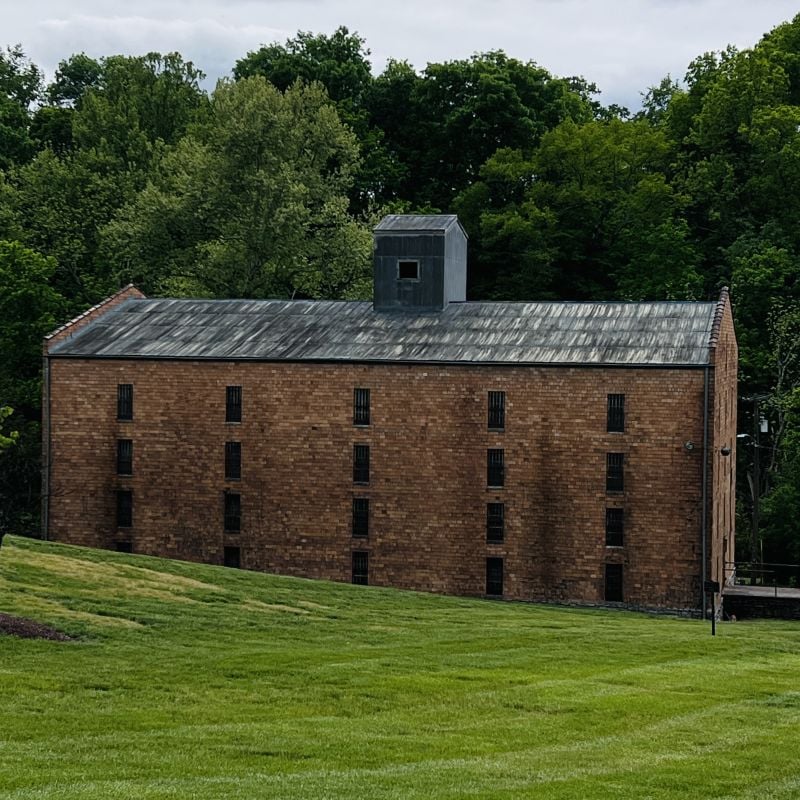A large, rectangular brick building with a metal roof and small windows sits on a grassy slope, surrounded by dense green trees under a cloudy sky.