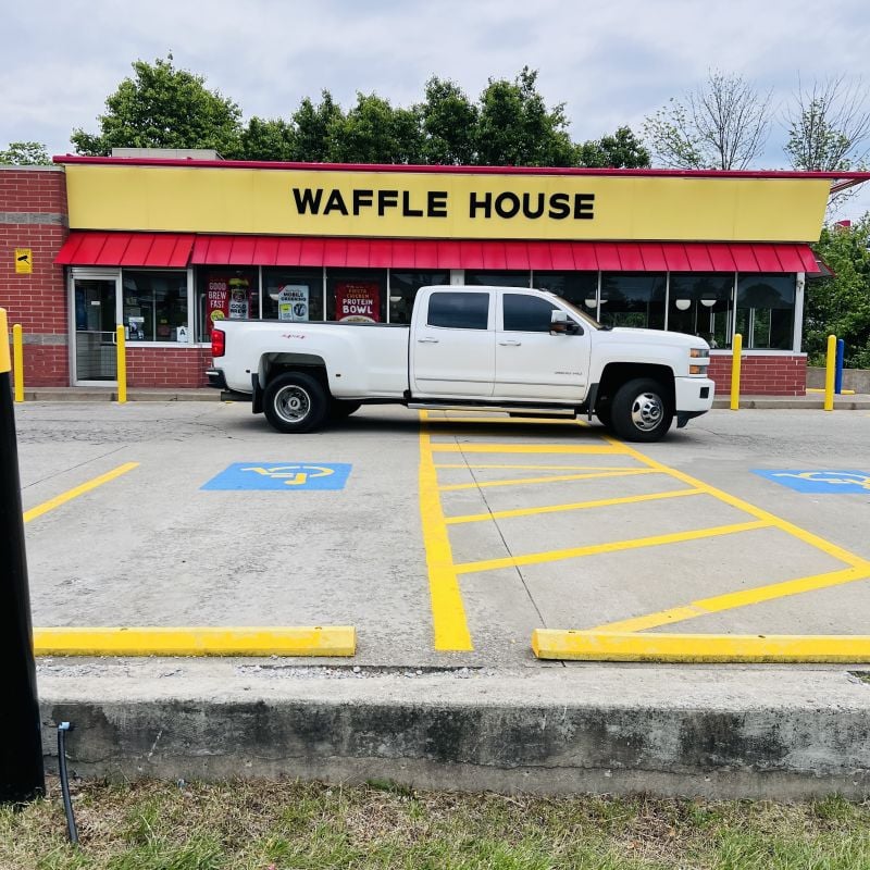 A white pickup truck is parked across two handicap spaces in front of a Waffle House restaurant with a yellow sign and red awning. Trees are visible behind the building.