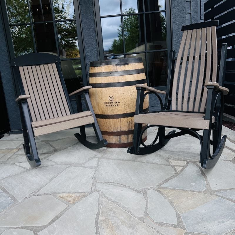 Two wooden rocking chairs with black frames sit on a stone patio beside a large wooden barrel, in front of windows reflecting trees and sky.
