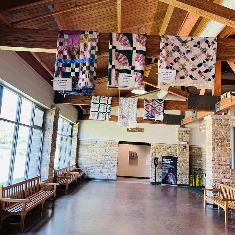 A bright hallway with stone walls, wooden benches, and colorful patchwork quilts hanging from the wooden-beamed ceiling. Large windows line the left side, and signs point to restrooms in the back.
