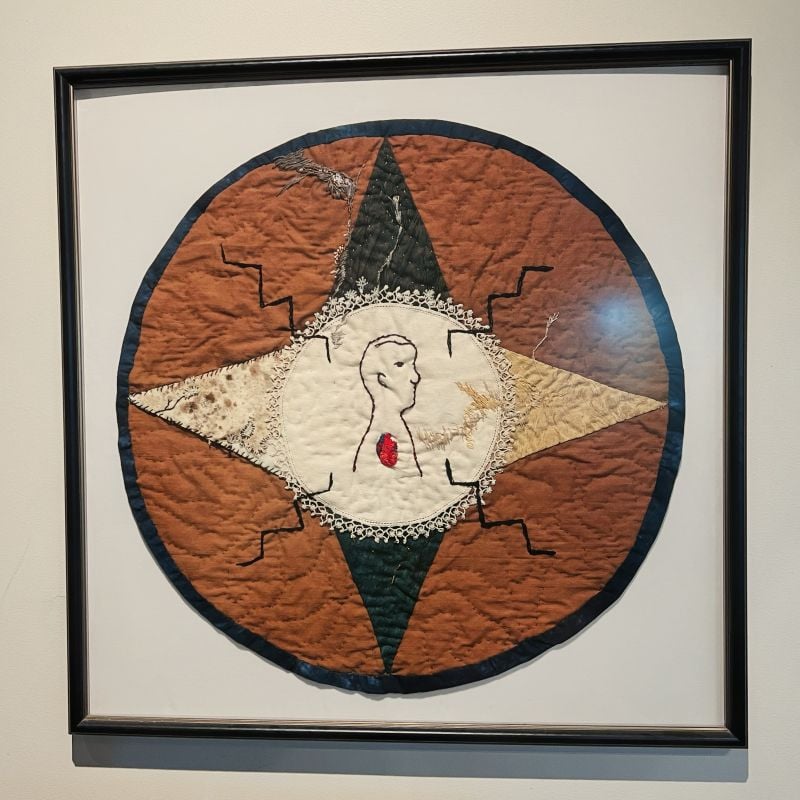 A framed textile artwork features a circular quilt with a central figure outlined in black and a red patch on the chest, surrounded by a star pattern in earthy tones of brown, orange, and green, displayed on a white background.