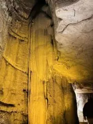 A tall, textured cave formation lit by warm yellow lighting rises from the cave floor to the ceiling, surrounded by rough, rocky walls inside a cavern.