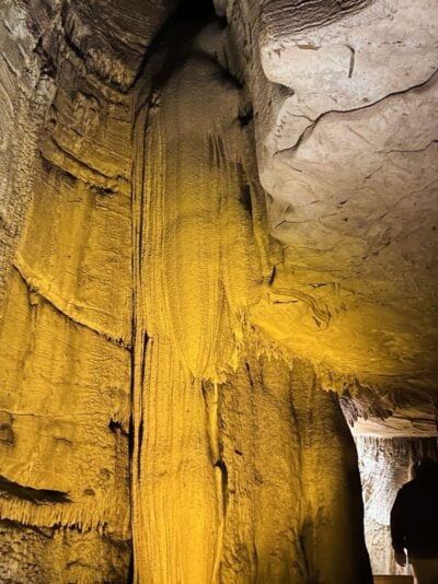 A tall, textured cave formation lit by warm yellow lighting rises from the cave floor to the ceiling, surrounded by rough, rocky walls inside a cavern.