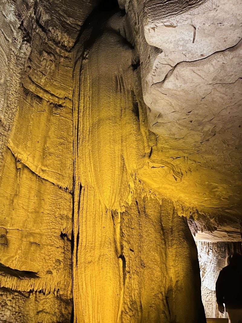 A tall, textured cave formation lit by warm yellow lighting rises from the cave floor to the ceiling, surrounded by rough, rocky walls inside a cavern.