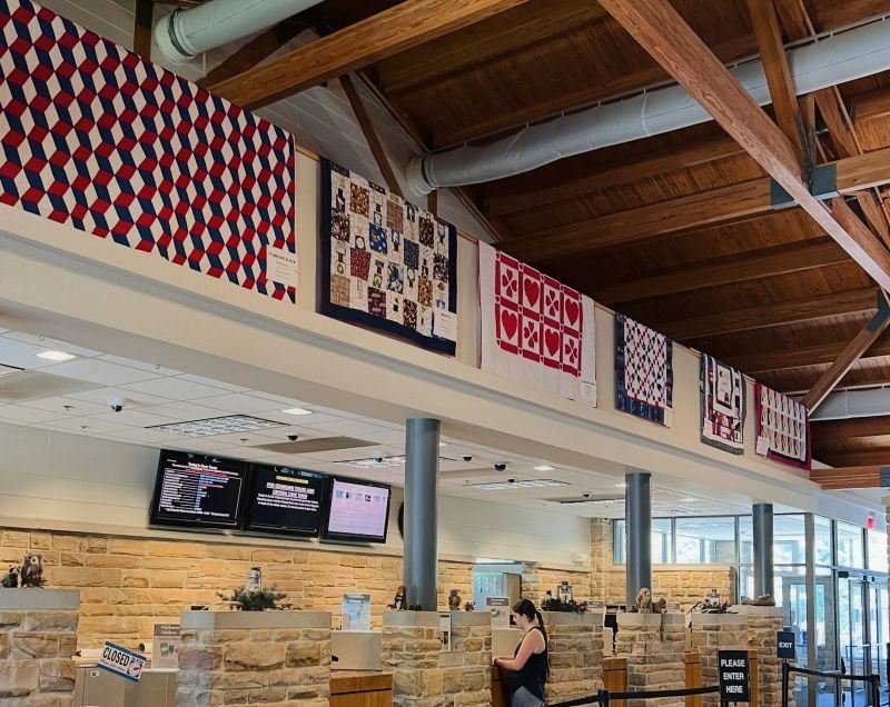 A woman stands in line inside a bank or office with stone walls and a wooden ceiling. Colorful quilts with geometric and heart patterns hang above the service counters. Two digital screens display information.