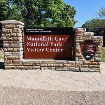 A stone sign reads Mammoth Cave National Park Visitor Center with the National Park Service logo on the right, set against a backdrop of green trees and blue sky.