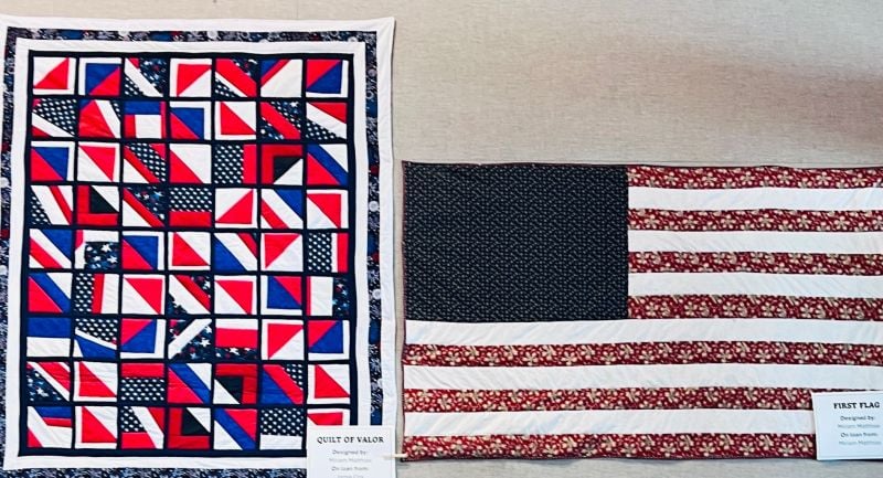 Two quilts are displayed side by side: one with a geometric red, white, and blue pattern, and the other resembling the American flag with stripes and a blue field of stars. Small placards are placed below each quilt.