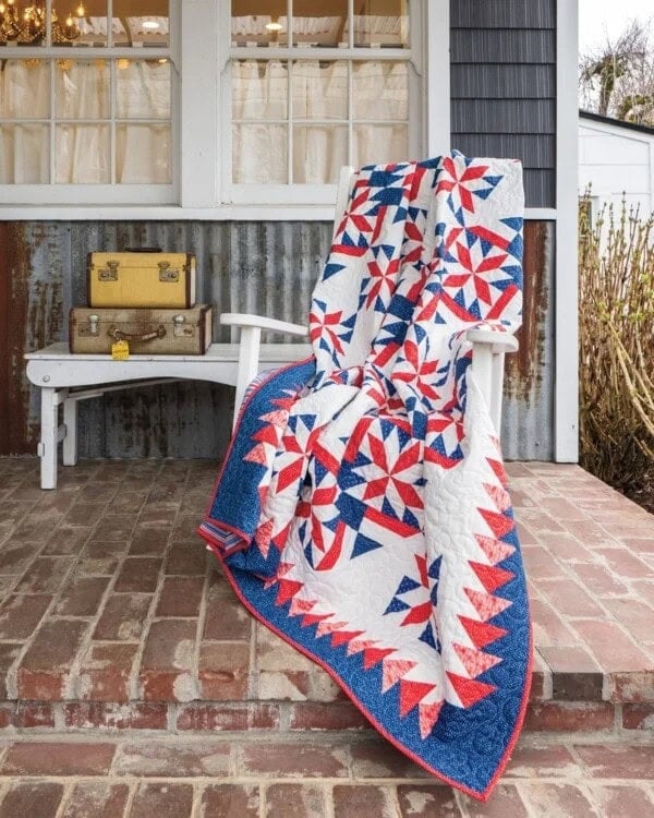 A white rocking chair on a brick porch holds a red, white, and blue patchwork quilt. Behind it, a white bench displays a vintage yellow suitcase and a brown trunk. Windows and rustic siding are in the background.