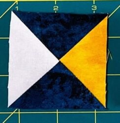 A Rising Star Quilt Block with four triangles: top left is white, top right is yellow, and the bottom two are dark blue, forming an hourglass pattern on a green cutting mat with a yellow grid.