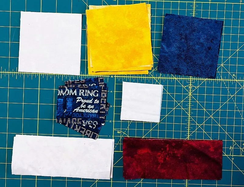 Assorted fabric squares and rectangles in white, yellow, blue, red, and patterned blue are arranged on a green cutting mat marked with a yellow grid.