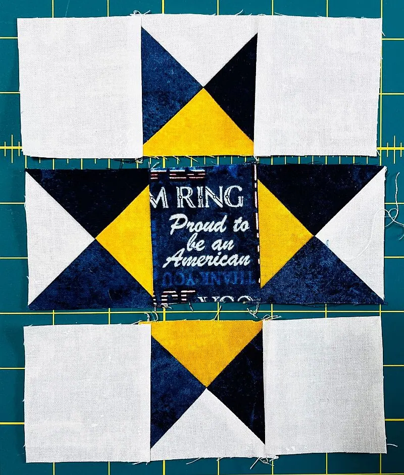 A quilt block layout with a center square of blue fabric with white text, surrounded by triangles of navy, yellow, and white fabric, arranged on a green cutting mat with yellow grid lines.