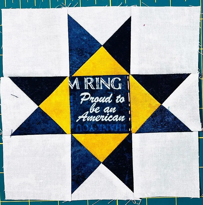A quilt block featuring a star pattern in navy blue and yellow on a white background, with the center reading, Proud to be an American and partial other text visible.