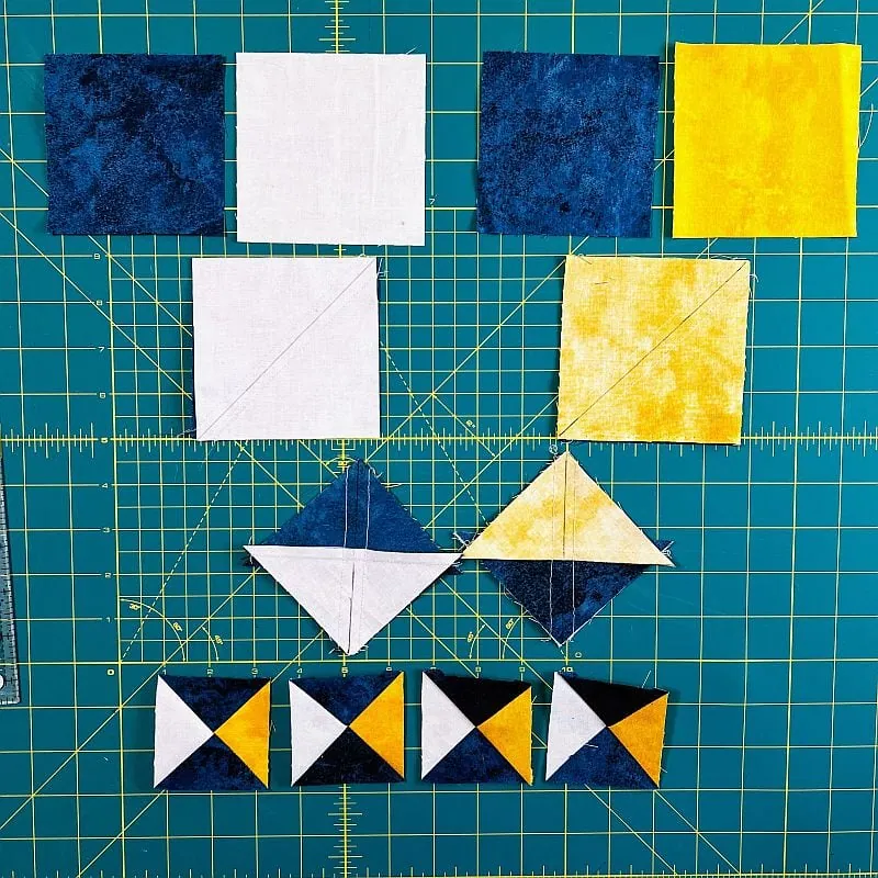 Squares of blue, white, and yellow fabric are arranged on a green cutting mat. Some squares are sewn into triangles, forming a row of Rising Star Quilt Block pieces with striking geometric patterns.