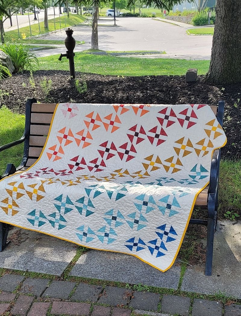 A colorful quilt with pinwheel patterns in red, orange, yellow, green, and blue is draped over a park bench on a sunny day, with grass and trees in the background.