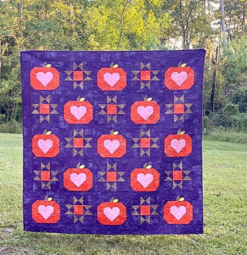 A quilt hangs outdoors, featuring alternating blocks of red pumpkins with pink hearts and star patterns on a purple background. Trees and grass are visible in the background.