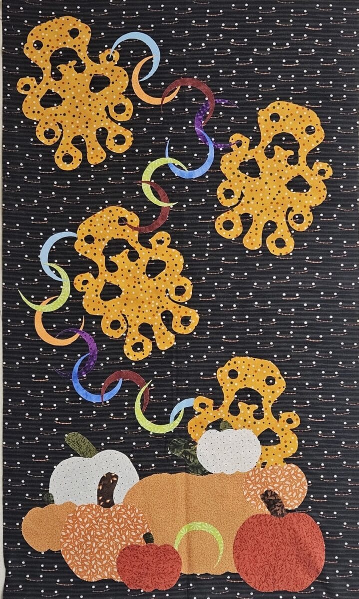 Quilted fabric art featuring orange octopus shapes, colorful streamers, and various pumpkins on a black background with small white dots, creating a festive autumn-themed scene.