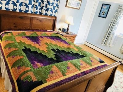 A wooden bed with a colorful quilt in shades of green, orange, yellow, and purple. The room has light blue walls, a lamp on a nightstand, framed art, and patterned curtains.