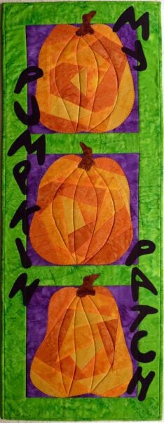 A quilted wall hanging with three patchwork pumpkins on purple squares, set against a green background. Black letters spell out Pumpkin Patch along the sides.
