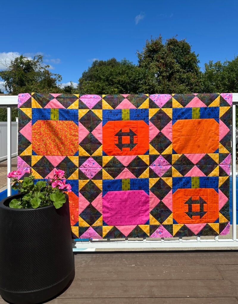 A colorful quilt with autumn pumpkin patterns hangs on a white fence outdoors near a large black planter with pink flowers. Blue sky and green trees are visible in the background.