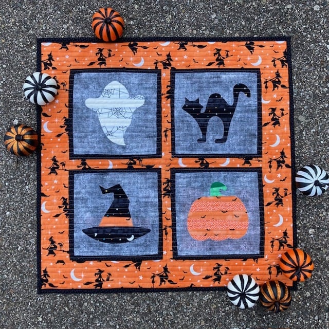 Halloween-themed quilt with four squares: a ghost, a cat, a witch hat, and a pumpkin. This autumn-inspired quilt features an orange and black pattern and is surrounded by small, striped decorative pumpkins on a textured surface. Perfect for cozying up as part of your seasonal Haunts Bundle decor.
